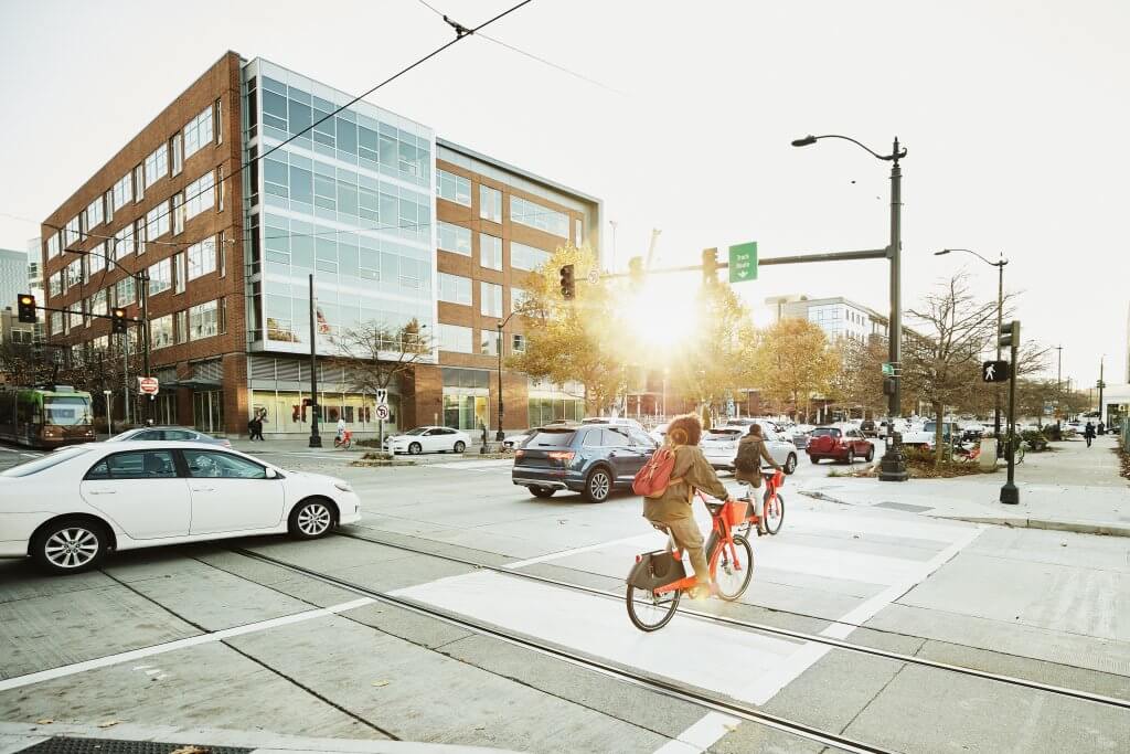 Pessoas andando de bicicleta no trânsito de uma cidade, carros circulam ao redor da ciclovia. Mobilidade urbana e o futuro das cidades