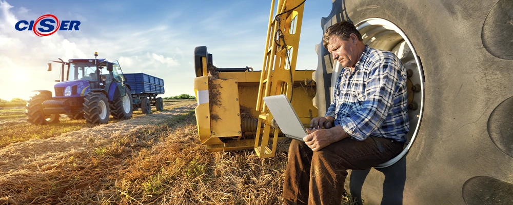 Homem sentado em roda de trator com notebook no colo, no campo